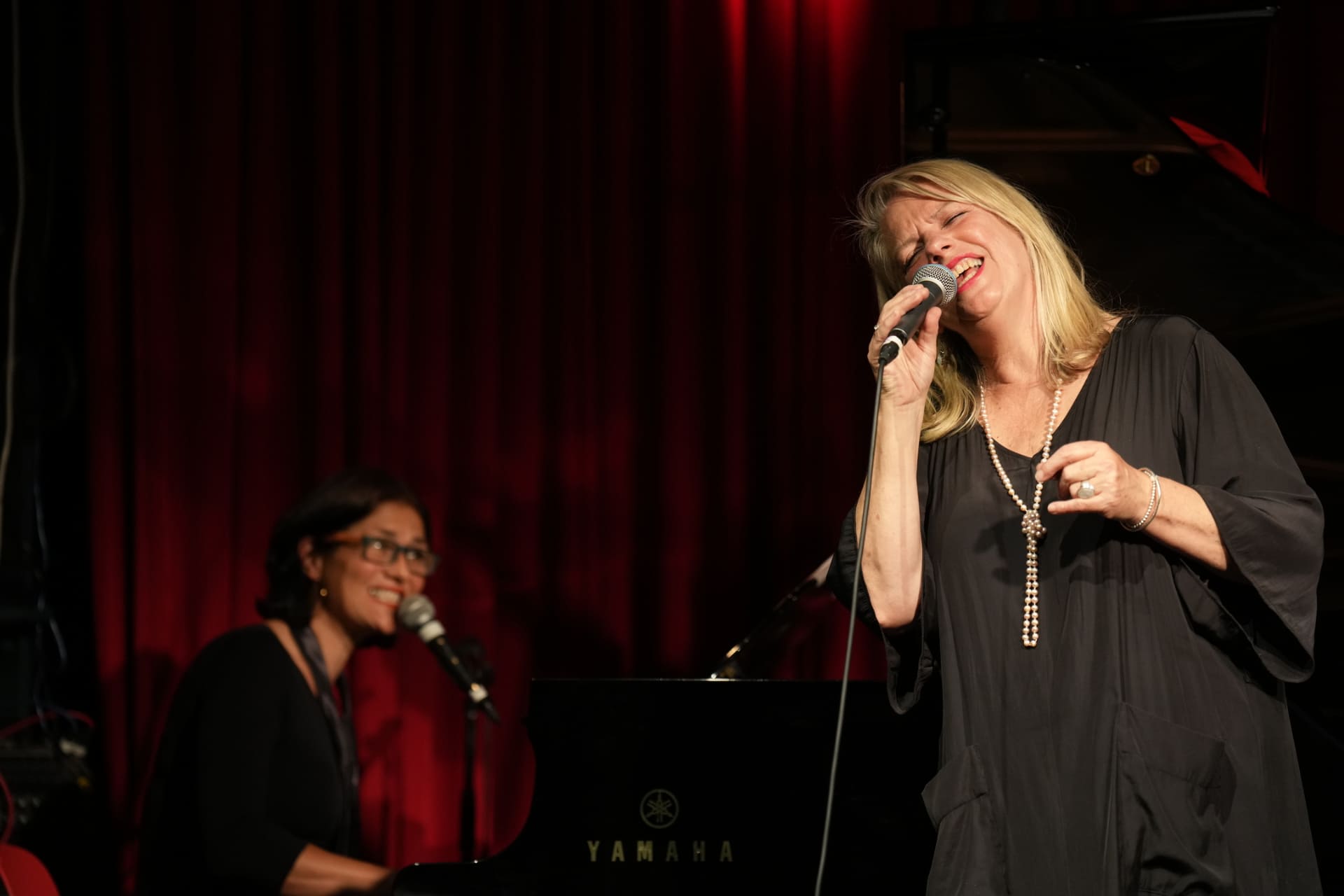 Rebecca Barnard on stage with a pianist, singing jazz. She is wearing a long black dress and pearl necklace