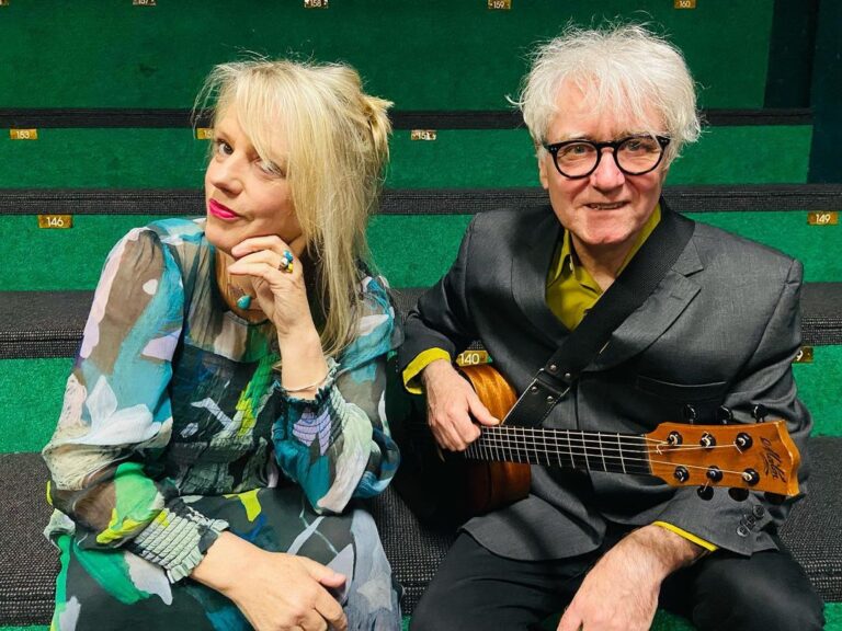 singer Rebecca Barnard and multi-instrumentalist Peter Farnan sitting in front of a green wall, Peter has a guitar on his lap, Rebecca is wearing a colourful long dress